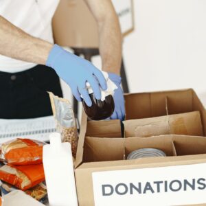 A volunteer with gloves packs canned goods and supplies in a donation box.